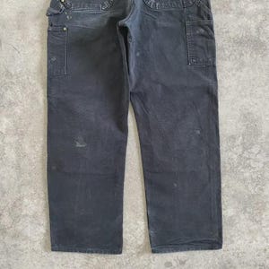 Puede incluir: Pantalones de trabajo gris oscuro con m&uacute;ltiples bolsillos y un lazo para martillo. Los pantalones muestran signos de desgaste, con algo de decoloraci&oacute;n y manchas. Los pantalones est&aacute;n hechos de un material duradero, adecuado para el trabajo o el uso informal.