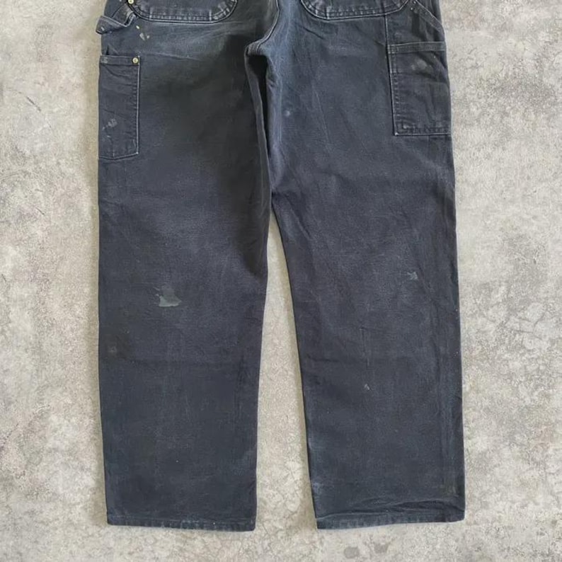 Puede incluir: Pantalones de trabajo gris oscuro con m&uacute;ltiples bolsillos y un lazo para martillo. Los pantalones muestran signos de desgaste, con algo de decoloraci&oacute;n y manchas. Los pantalones est&aacute;n hechos de un material duradero, adecuado para el trabajo o el uso informal.