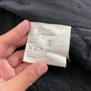 May include: Close-up of a clothing tag with Japanese text and care symbols. The tag is white with black text and includes details like "WD-P025", fabric composition, and size "S". The tag is held against a dark gray fabric.