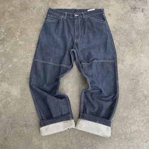 Puede incluir: Pantalones vaqueros azul oscuro con costuras blancas y dobladillos. Los pantalones tienen un diseño clásico de cinco bolsillos y cierre de botón. La mezclilla parece ser de peso medio, adecuada para varias estaciones. Los pantalones están colocados sobre una superficie de hormigón.