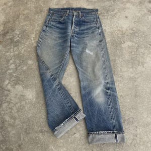 May include: Faded blue denim jeans with a button fly and distressed details. The jeans have a classic five-pocket design and are cuffed at the ankles, showcasing a lighter wash on the inside of the cuff. The jeans appear worn and well-loved.
