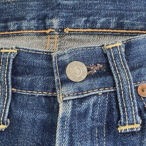 May include: Close-up of the button and buttonhole area of blue denim jeans. The jeans have a metal button with the text "McCOY'S" around the edge. The stitching is yellow and the denim is faded.