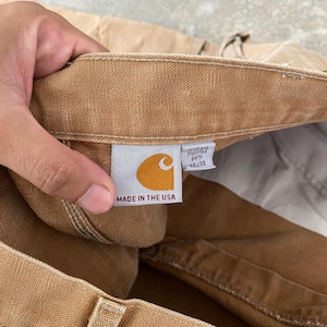 K&ouml;nnte beinhalten: Nahaufnahme einer beigefarbenen Arbeitshose mit einem Carhartt-Etikett. Das Etikett zeigt ein goldenes Logo und den Text "MADE IN THE USA". Die Hose hat die Gr&ouml;&szlig;e 44x30. Das Etikett sagt auch "parts" und "imported".
