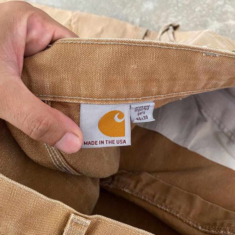 K&ouml;nnte beinhalten: Nahaufnahme einer beigefarbenen Arbeitshose mit einem Carhartt-Etikett. Das Etikett zeigt ein goldenes Logo und den Text "MADE IN THE USA". Die Hose hat die Gr&ouml;&szlig;e 44x30. Das Etikett sagt auch "parts" und "imported".