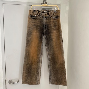 May include: Wide-leg jeans in a brown and black acid wash. The denim pants are hanging on a hanger against a white door. The jeans have a button and zipper closure, with pockets on the front and back.
