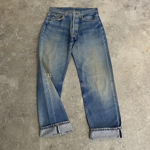 Könnte beinhalten: Eine mittelblaue Jeans aus Denim mit Knopfleiste, fünf Taschen und einer sichtbaren Webkante an den umgeschlagenen Säumen. Der Stoff weist natürliche Verblassungen und eine kleine abgenutzte Stelle am rechten Oberschenkel auf.