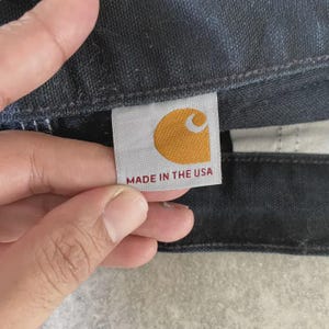 Puede incluir: Primer plano de una etiqueta Carhartt, con un logotipo de media luna amarillo dorado sobre fondo blanco. El texto "MADE IN THE USA" est&aacute; impreso en rojo. La etiqueta est&aacute; adherida a tela vaquera oscura.