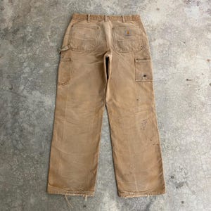 K&ouml;nnte beinhalten: Beige Arbeitshose mit mehreren Taschen, einschlie&szlig;lich einer Hammerschlaufe. Die Hose weist Gebrauchsspuren auf, mit sichtbaren Flecken und ausgefransten Kanten. Ein kleines Logo-Patch befindet sich auf der Ges&auml;&szlig;tasche. Die Hose ist aus strapazierf&auml;higem Stoff gefertigt.