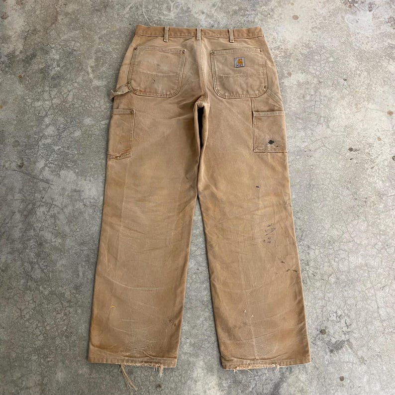 K&ouml;nnte beinhalten: Beige Arbeitshose mit mehreren Taschen, einschlie&szlig;lich einer Hammerschlaufe. Die Hose weist Gebrauchsspuren auf, mit sichtbaren Flecken und ausgefransten Kanten. Ein kleines Logo-Patch befindet sich auf der Ges&auml;&szlig;tasche. Die Hose ist aus strapazierf&auml;higem Stoff gefertigt.