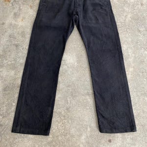 May include: Black denim pants with a classic five-pocket design. The pants have a straight leg cut and a button and zipper closure. The fabric appears slightly worn, giving them a vintage look. The pants are laid flat on a concrete surface.