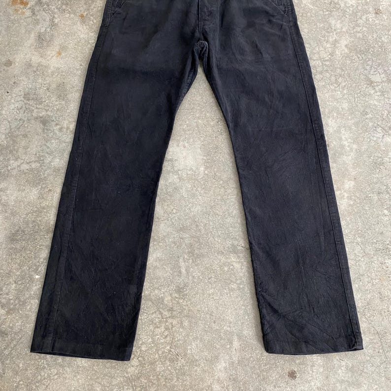 May include: Black denim pants with a classic five-pocket design. The pants have a straight leg cut and a button and zipper closure. The fabric appears slightly worn, giving them a vintage look. The pants are laid flat on a concrete surface.