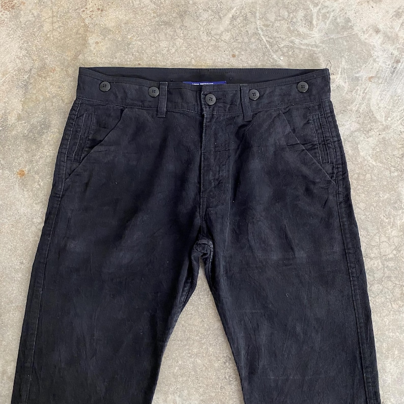 May include: Black corduroy pants with button details. The pants have a classic five-pocket design and a button fly. The fabric has a subtle vertical ribbing. The pants are a solid black color and appear to be well-worn.