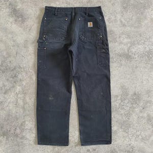 Puede incluir: Pantalones de trabajo gris oscuro con m&uacute;ltiples bolsillos, incluyendo dos bolsillos traseros y bolsillos laterales. Los pantalones tienen un peque&ntilde;o parche cuadrado con el logo en el bolsillo trasero. Los pantalones son de material duradero y muestran signos de desgaste.