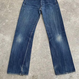 May include: Faded blue denim jeans with a button fly. The jeans show wear and tear, with lighter patches on the thighs and knees. The straight-leg style is visible, and the jeans are laid flat.