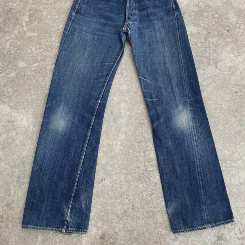 May include: Faded blue denim jeans with a button fly. The jeans show wear and tear, with lighter patches on the thighs and knees. The straight-leg style is visible, and the jeans are laid flat.