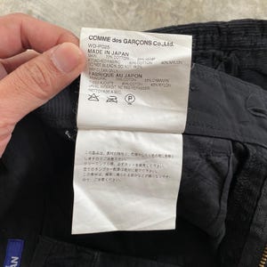 May include: Close-up of a tag from Comme des Gar&ccedil;ons Co., Ltd. The tag is white with black text, listing fabric composition: 70% cotton, 30% hemp, 60% cotton, and 40% nylon. The tag also includes washing instructions.