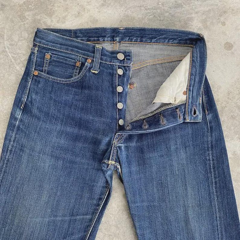 May include: Blue denim jeans with a button fly. The jeans have a classic five-pocket design, with rivets at the corners of the pockets. The denim has a faded appearance, suggesting wear. The jeans are unbuttoned, showing the interior.