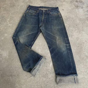 May include: Faded blue denim jeans with a button fly and folded cuffs. The jeans show signs of wear and fading, with visible creases and a classic five-pocket design. The denim has a vintage, worn-in look.