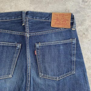 May include: Close-up of the back of blue denim jeans. The jeans have two back pockets and a small red tag on one pocket. A brown leather patch with the text "JOE McCOY & CO." is visible at the waistband.
