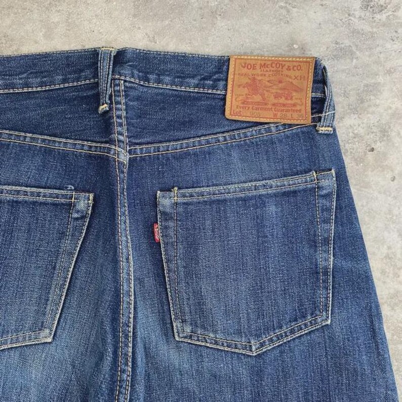 May include: Close-up of the back of blue denim jeans. The jeans have two back pockets and a small red tag on one pocket. A brown leather patch with the text "JOE McCOY & CO." is visible at the waistband.