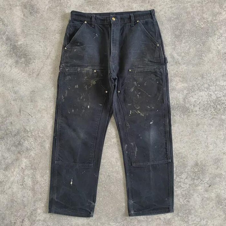 Puede incluir: Pantalones de trabajo de color gris oscuro con m&uacute;ltiples bolsillos y rodillas reforzadas. Los pantalones muestran signos de desgaste con salpicaduras de pintura y un aspecto desgastado. Los pantalones de trabajo est&aacute;n hechos de un material duradero.