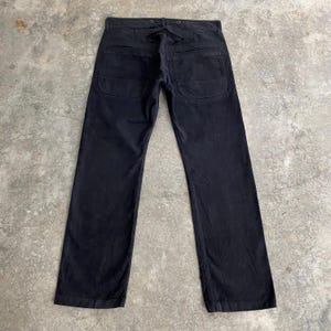 May include: Black work pants with two large back pockets and a tie closure at the waist. The pants have a straight leg design and are made of a durable fabric. The pants are laid flat on a concrete surface.