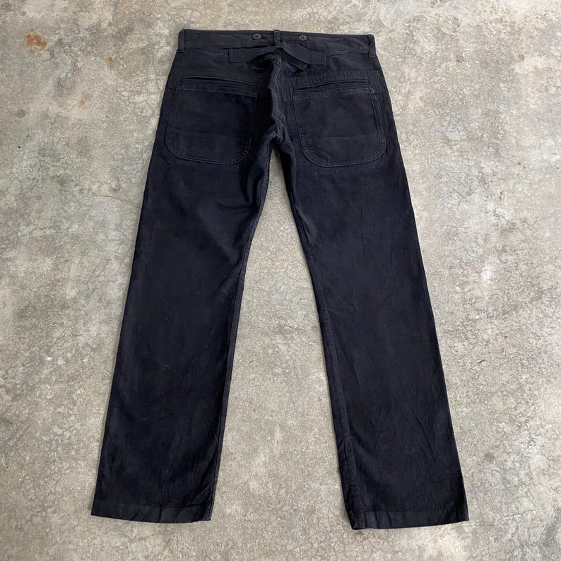 May include: Black work pants with two large back pockets and a tie closure at the waist. The pants have a straight leg design and are made of a durable fabric. The pants are laid flat on a concrete surface.