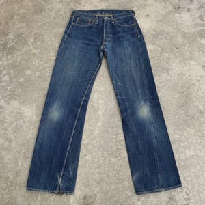 May include: Blue denim jeans with a classic five-pocket design. The jeans have a faded wash and subtle wear marks, giving them a vintage look. The straight-leg cut and button fly are visible. The jeans are laid flat on a concrete surface.