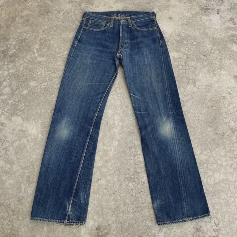 May include: Blue denim jeans with a classic five-pocket design. The jeans have a faded wash and subtle wear marks, giving them a vintage look. The straight-leg cut and button fly are visible. The jeans are laid flat on a concrete surface.