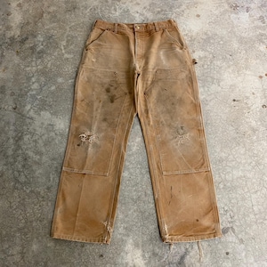 K&ouml;nnte beinhalten: Eine abgenutzte, beige Arbeitshose mit verst&auml;rkten Kniepatches und mehreren Taschen. Die Hose weist Gebrauchsspuren auf, darunter Flecken und abgenutzte Stellen. Die Hose hat einen Knopf- und Rei&szlig;verschluss am Bund.