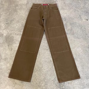 May include: Brown denim trousers with large square patches on the knees. The trousers have a wide leg design and classic five-pocket styling. The trousers are made of a durable denim material and feature a button and zip closure.