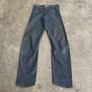 May include: Faded blue denim jeans with a classic five-pocket design. The jeans have a straight leg cut and show signs of wear, giving them a vintage look. The jeans are laid flat on a concrete surface.