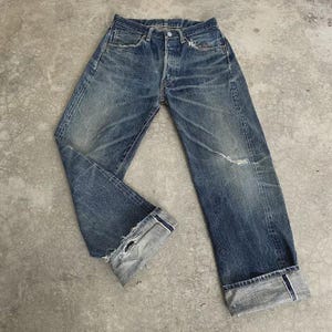 May include: Faded blue denim jeans with a button fly and distressed details. The jeans have a worn appearance with visible wear marks and a small tear on the knee. The cuffs are rolled up, revealing a lighter shade of denim.