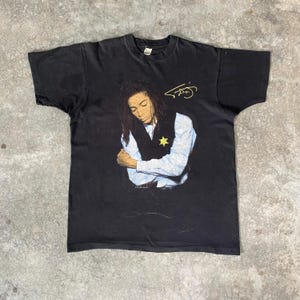 May include: Black t-shirt featuring a graphic of a person with dreadlocks, wearing a vest and button-down shirt. A yellow signature is above the graphic. The shirt is vintage and shows signs of wear.