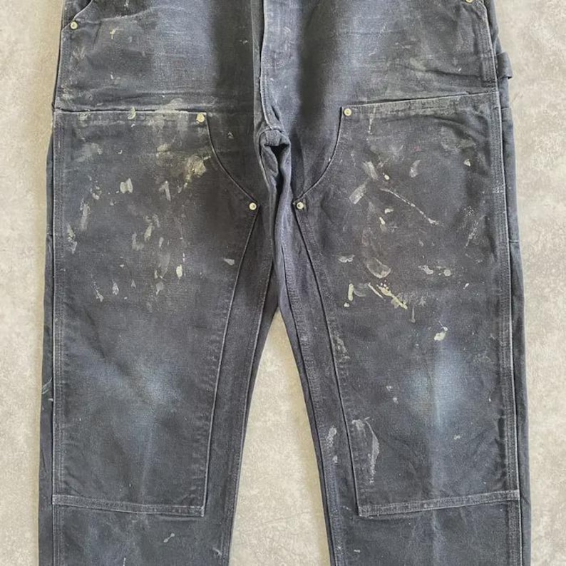 Puede incluir: Pantalones de trabajo gris oscuro con m&uacute;ltiples bolsillos y detalles de remaches. Los pantalones tienen salpicaduras de pintura en la parte delantera, lo que sugiere su uso en un taller o estudio de arte. Los pantalones est&aacute;n hechos de un material duradero.