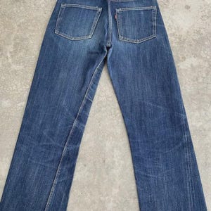 May include: Back view of blue denim jeans with two back pockets and a red tag. The jeans have a classic straight-leg fit and show signs of wear and fading, giving them a vintage look.