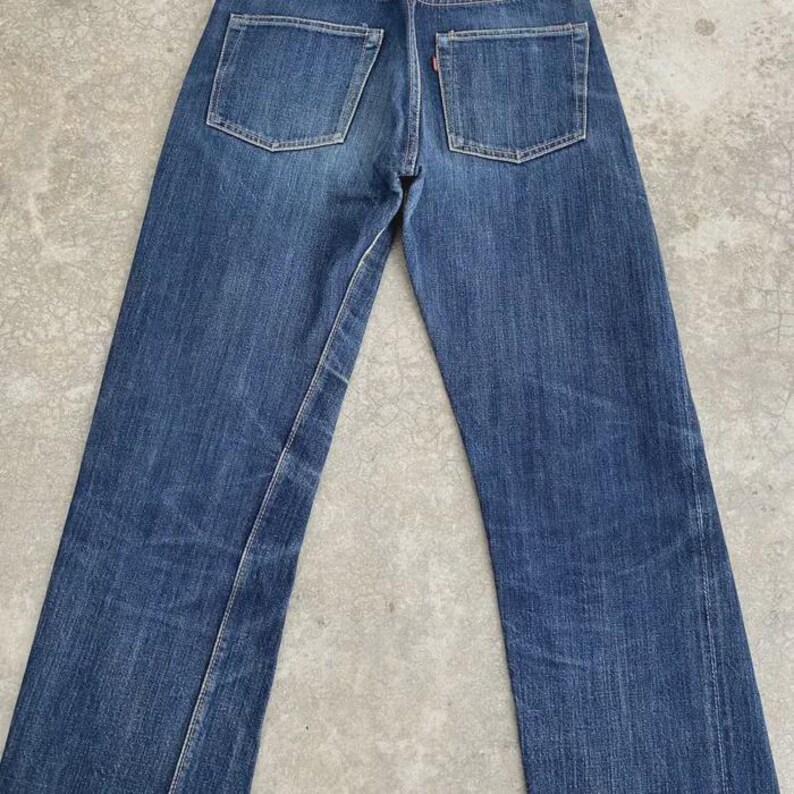 May include: Back view of blue denim jeans with two back pockets and a red tag. The jeans have a classic straight-leg fit and show signs of wear and fading, giving them a vintage look.