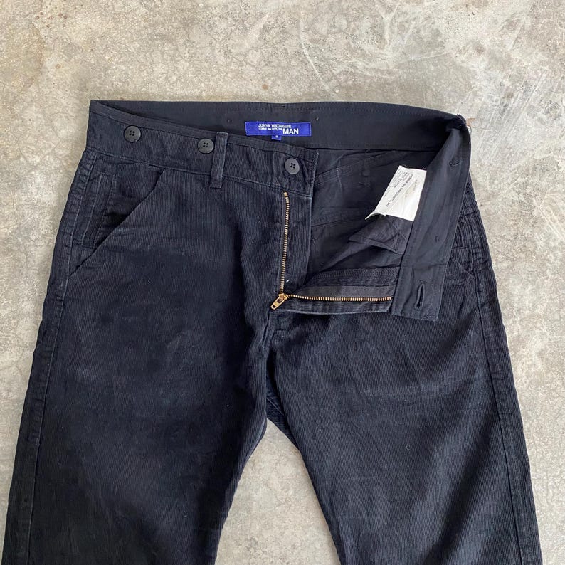 May include: Black corduroy pants with a button and zipper closure. The pants have a classic five-pocket design and a straight leg fit. The label reads "JUNYA WATANABE MAN".