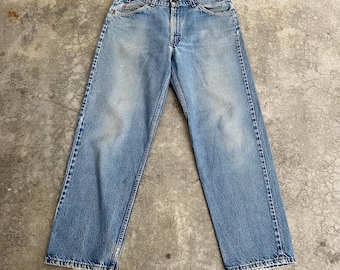 May include: Light blue denim jeans with a classic five-pocket design. The jeans show signs of wear and fading, with a straight leg cut and frayed hems. The jeans are laid flat on a concrete surface.