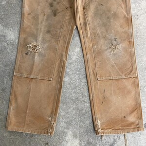 K&ouml;nnte beinhalten: Nahaufnahme einer abgenutzten, beigefarbenen Arbeitshose mit sichtbaren Gebrauchsspuren. Die Hose hat mehrere Taschen, verst&auml;rkte Knie und besch&auml;digte Bereiche, einschlie&szlig;lich L&ouml;chern und Flecken. Die Gesamt&auml;sthetik deutet auf ein Vintage- oder stark genutztes Kleidungsst&uuml;ck hin.