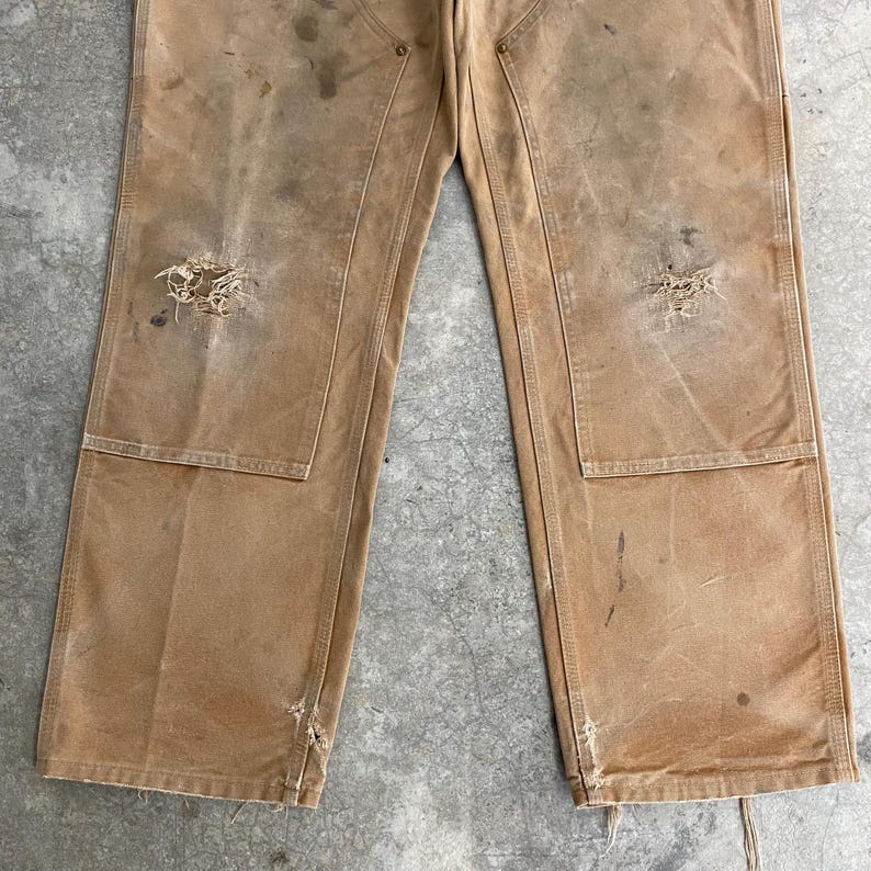 K&ouml;nnte beinhalten: Nahaufnahme einer abgenutzten, beigefarbenen Arbeitshose mit sichtbaren Gebrauchsspuren. Die Hose hat mehrere Taschen, verst&auml;rkte Knie und besch&auml;digte Bereiche, einschlie&szlig;lich L&ouml;chern und Flecken. Die Gesamt&auml;sthetik deutet auf ein Vintage- oder stark genutztes Kleidungsst&uuml;ck hin.