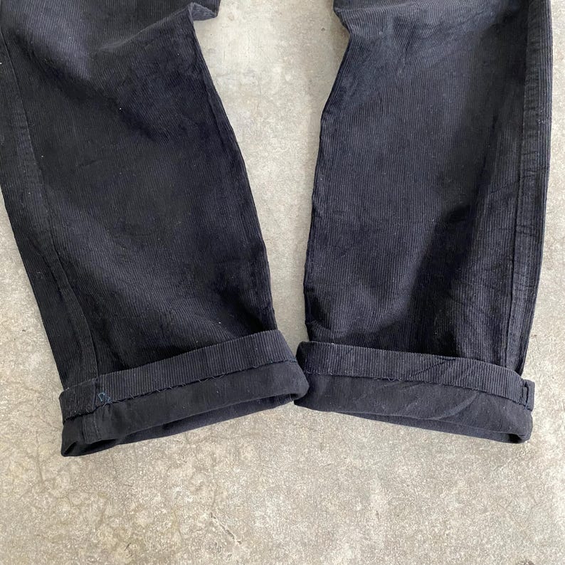 May include: Close-up of black corduroy pants with cuffed hems. The pants have a classic, straight-leg design and a textured, ribbed fabric. The cuffs are neatly folded, showcasing the attention to detail in the garment's construction.