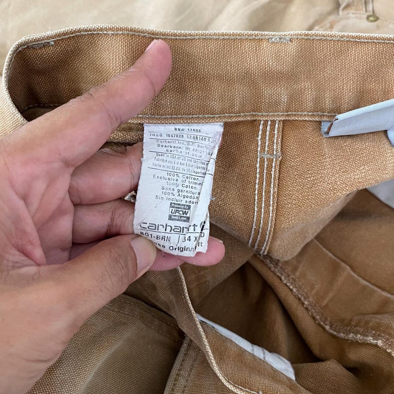 K&ouml;nnte beinhalten: Nahaufnahme einer beigefarbenen Carhartt-Arbeitshose. Das Etikett lautet "Carhartt" und enth&auml;lt Details zur Materialzusammensetzung, einschlie&szlig;lich 100 % Baumwolle. Die Hose hat die Gr&ouml;&szlig;e 34X.