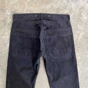 May include: Black corduroy pants with a unique design. The pants feature two large back pockets, a belt loop, and button details. The fabric has a ribbed texture.