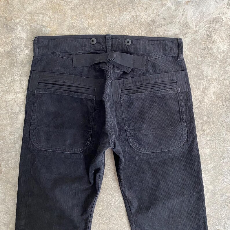 May include: Black corduroy pants with a unique design. The pants feature two large back pockets, a belt loop, and button details. The fabric has a ribbed texture.