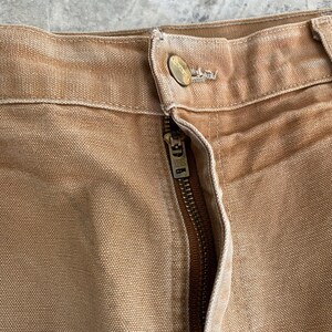 K&ouml;nnte beinhalten: Nahaufnahme einer beigefarbenen Arbeitshose mit sichtbarem Rei&szlig;verschluss und Knopfverschluss. Die Hose besteht aus einem strapazierf&auml;higen, strukturierten Stoff. Der Rei&szlig;verschluss ist messingfarben und der Knopf hat ein Logo.