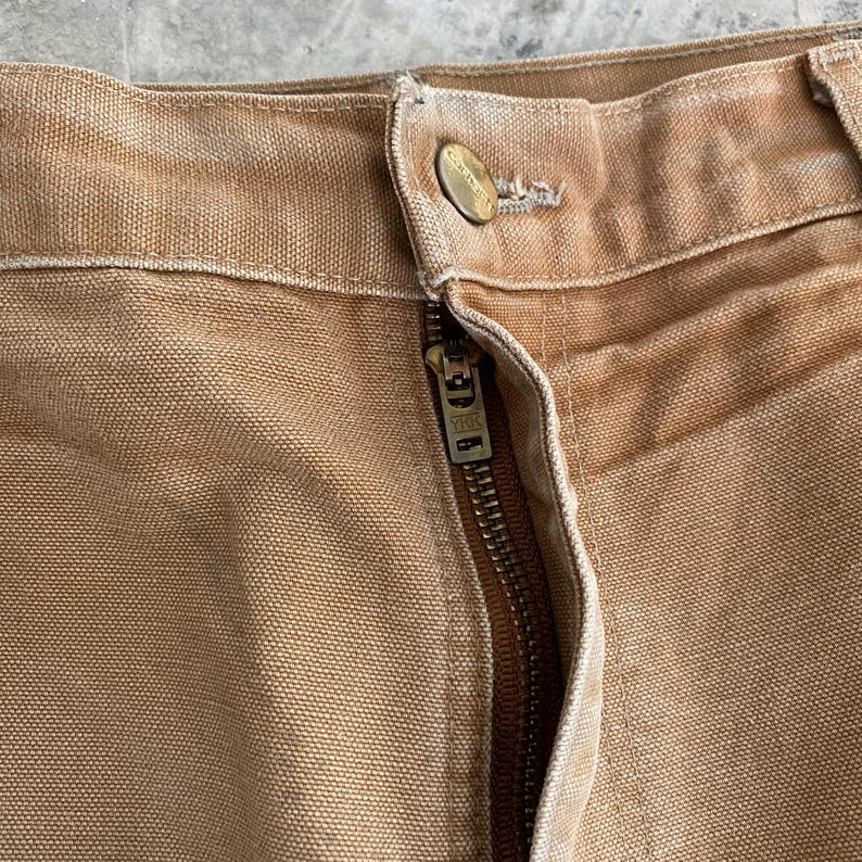 K&ouml;nnte beinhalten: Nahaufnahme einer beigefarbenen Arbeitshose mit sichtbarem Rei&szlig;verschluss und Knopfverschluss. Die Hose besteht aus einem strapazierf&auml;higen, strukturierten Stoff. Der Rei&szlig;verschluss ist messingfarben und der Knopf hat ein Logo.