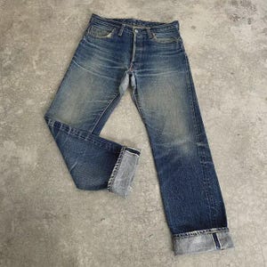 May include: Faded blue denim jeans with a button fly and folded cuffs. The jeans have a classic five-pocket design and show signs of wear, with lighter areas on the thighs and knees. The cuffs are folded up, revealing the selvedge edge.