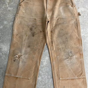 K&ouml;nnte beinhalten: Beige Arbeitshose mit mehreren Taschen und abgenutzten Details. Die Hose weist Gebrauchsspuren auf, darunter Flecken und Risse, was ihr eine Vintage-&Auml;sthetik verleiht. Die Hose hat ein klassisches Arbeitskleidungsdesign.