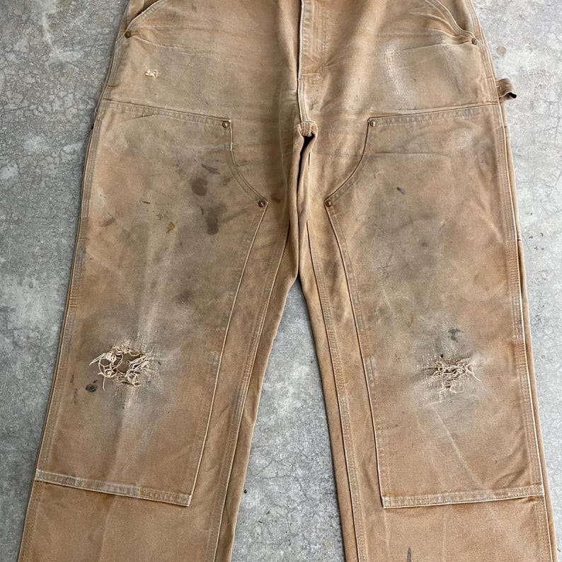 K&ouml;nnte beinhalten: Beige Arbeitshose mit mehreren Taschen und abgenutzten Details. Die Hose weist Gebrauchsspuren auf, darunter Flecken und Risse, was ihr eine Vintage-&Auml;sthetik verleiht. Die Hose hat ein klassisches Arbeitskleidungsdesign.